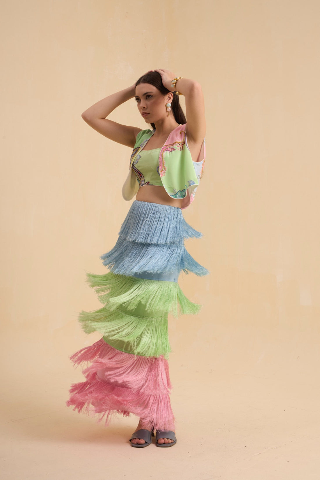 A woman modeling a colorful tiered ruffle costume set consisting of a light green crop top with pink and blue ruffle sleeves, a light blue ruffled skirt, a light green ruffled skirt, and a pink ruffled skirt, against a solid beige background.